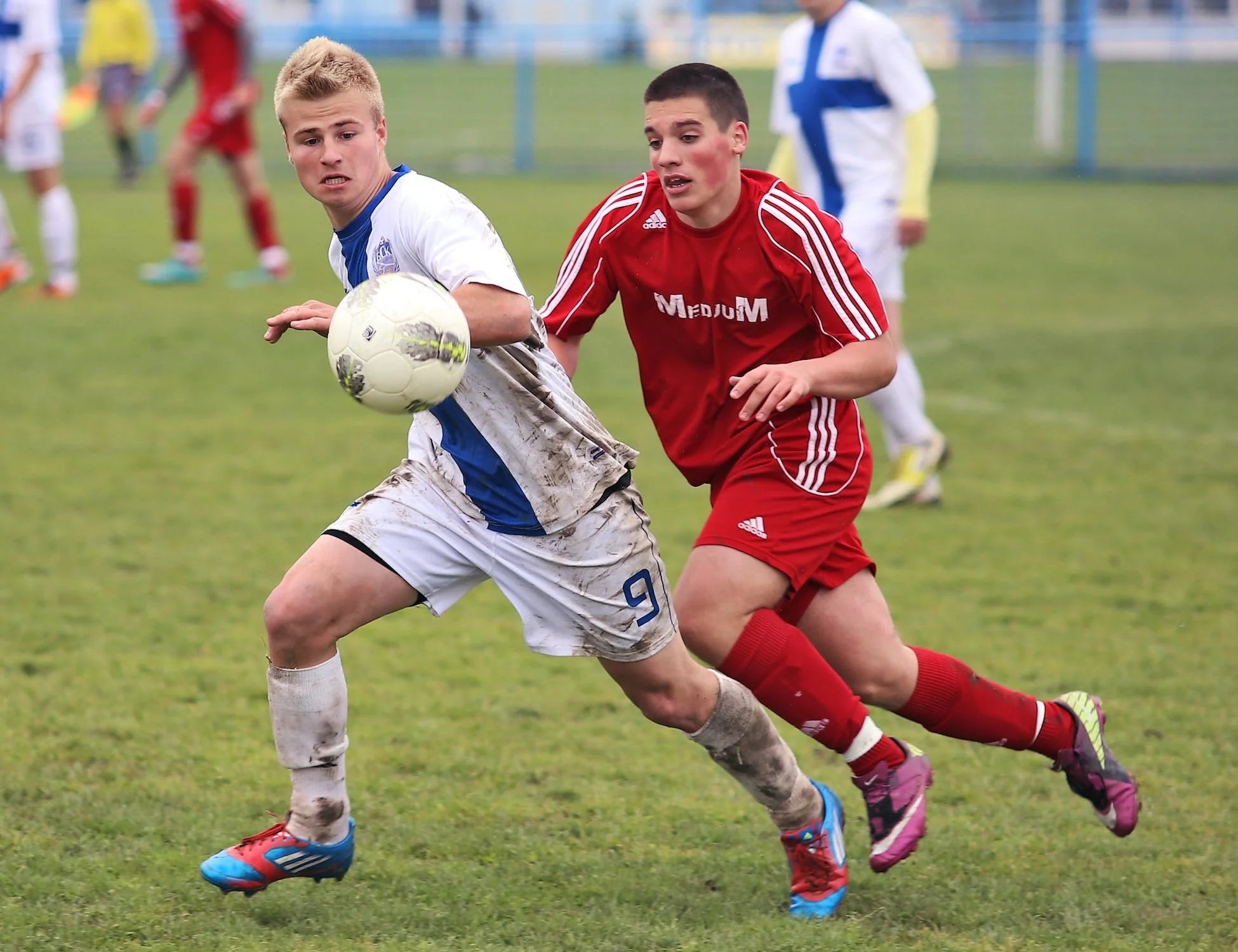 Two soccer players in action on the field, one in white and blue, the other in red.
