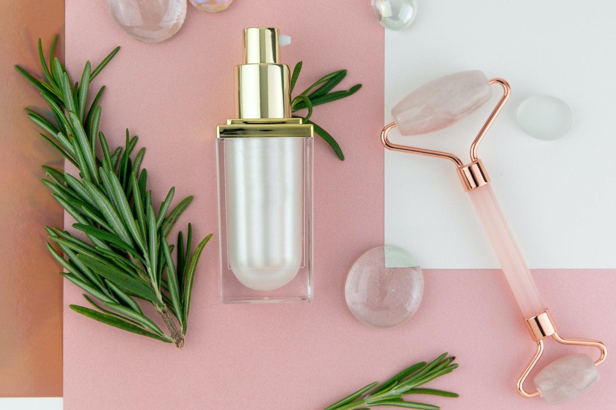 Rose quartz roller, glass serum bottle, and green rosemary sprigs on pink background.