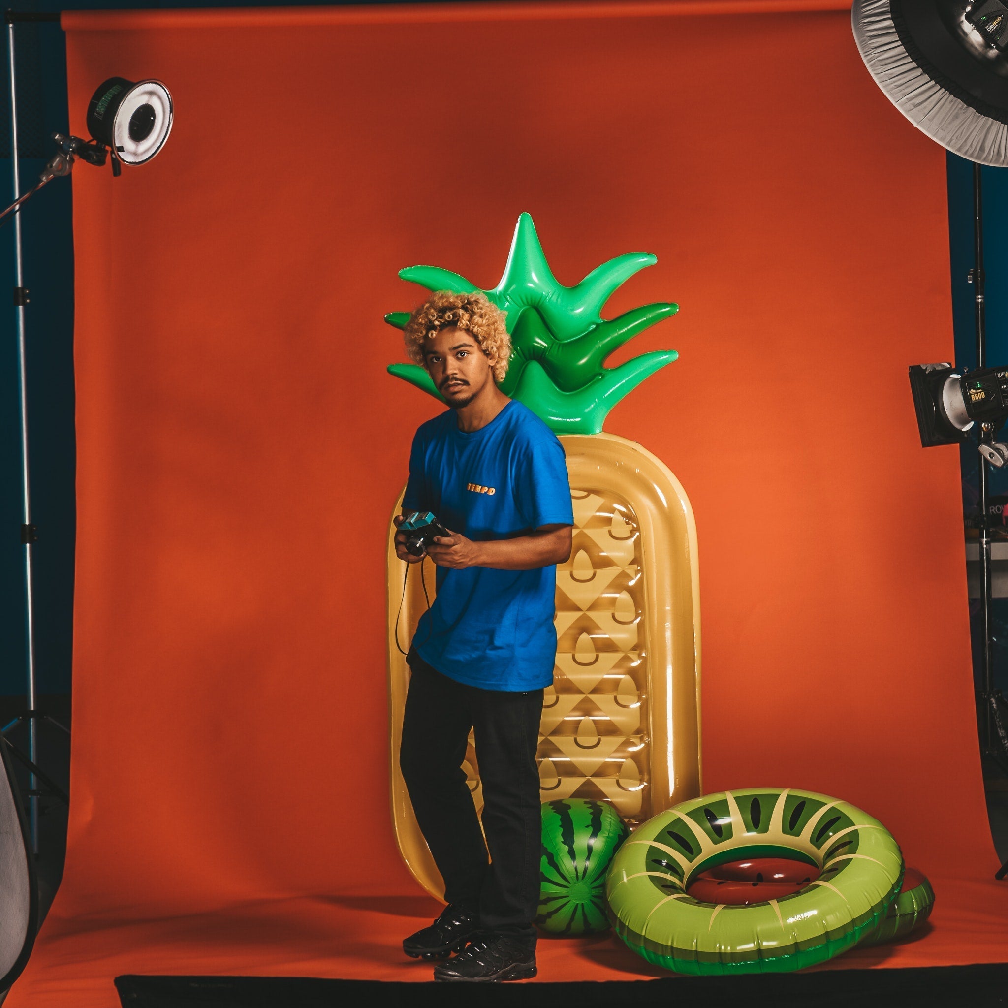 A person in a blue shirt stands with a game controller in front of bright orange backdrop with inflatable fruit.