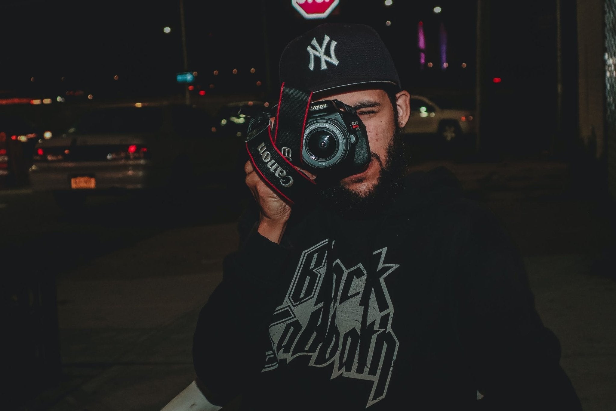 Person in dark hoodie holding a Canon camera, pointing it toward the viewer.