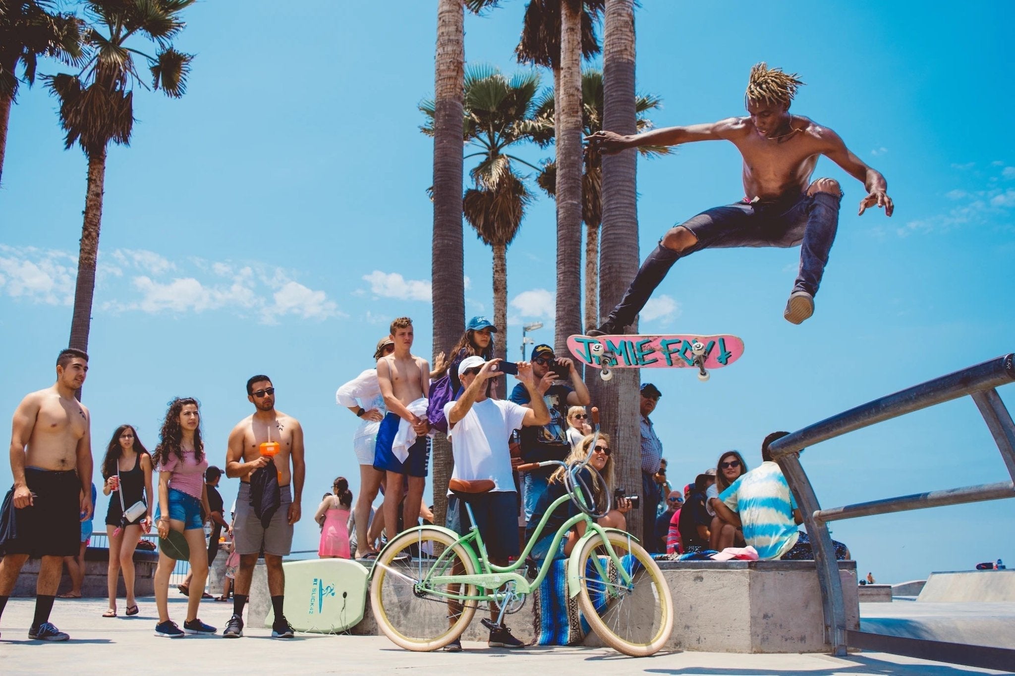 Outdoor skateboarding scene with a skateboarder performing a trick, spectators and palm trees in background.