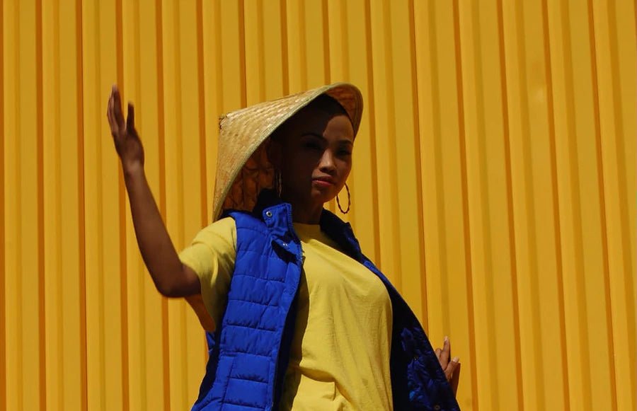 Woman in a blue vest and yellow shirt, wearing a straw hat, against a bright yellow background.