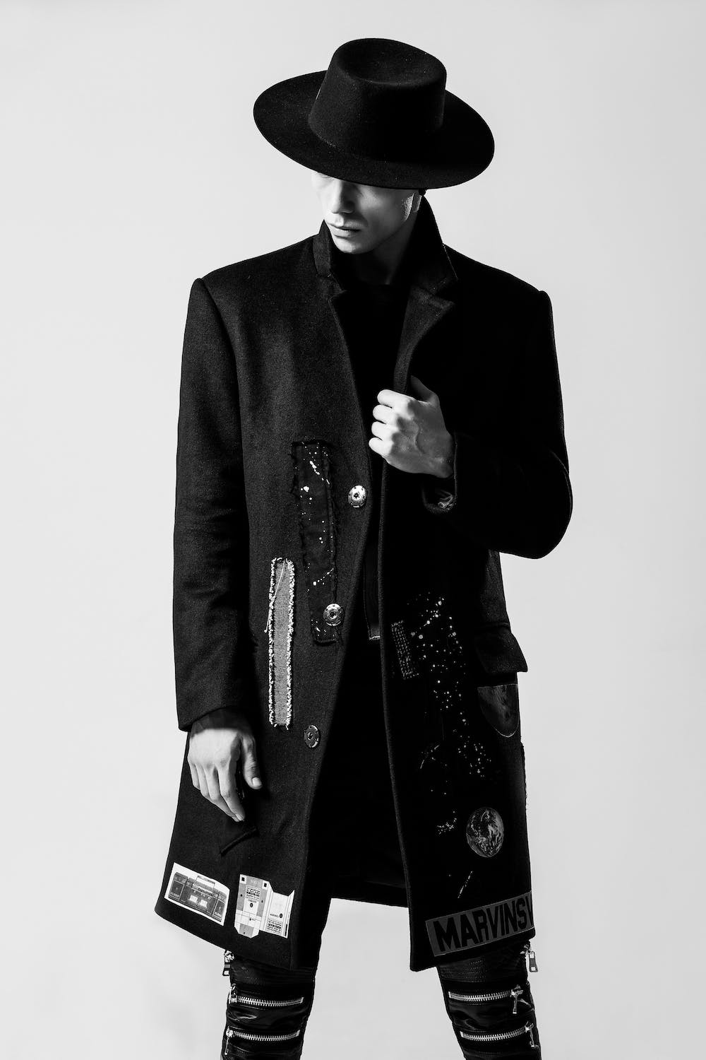 Black and white image of a model in a stylish coat and hat, striking a pose against a plain backdrop.