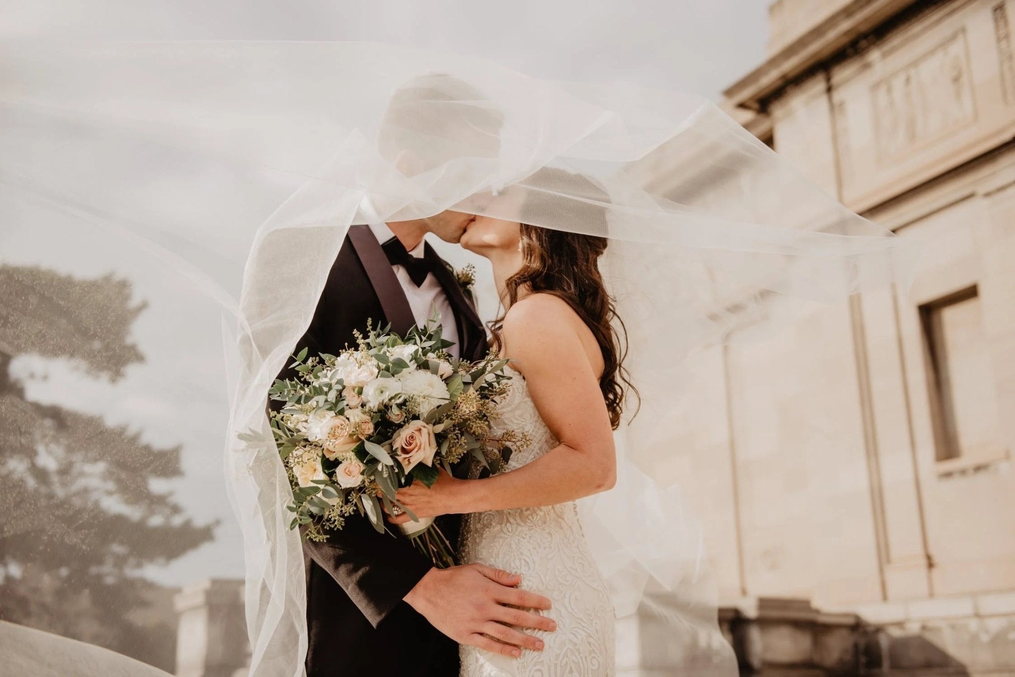 Couple embracing under a veil, holding a bouquet, with a historic building in the background.