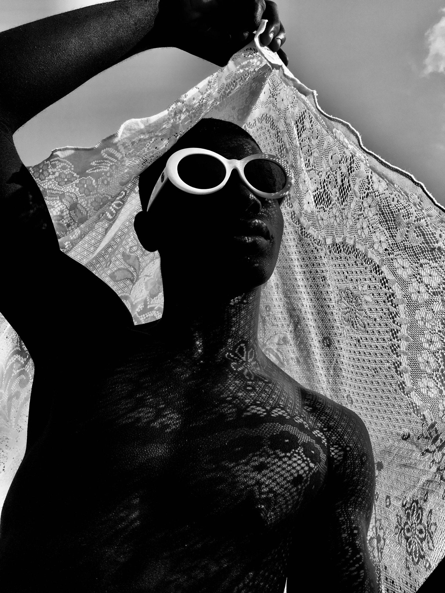 Black and white portrait of a person holding a lace cloth, wearing white round sunglasses.