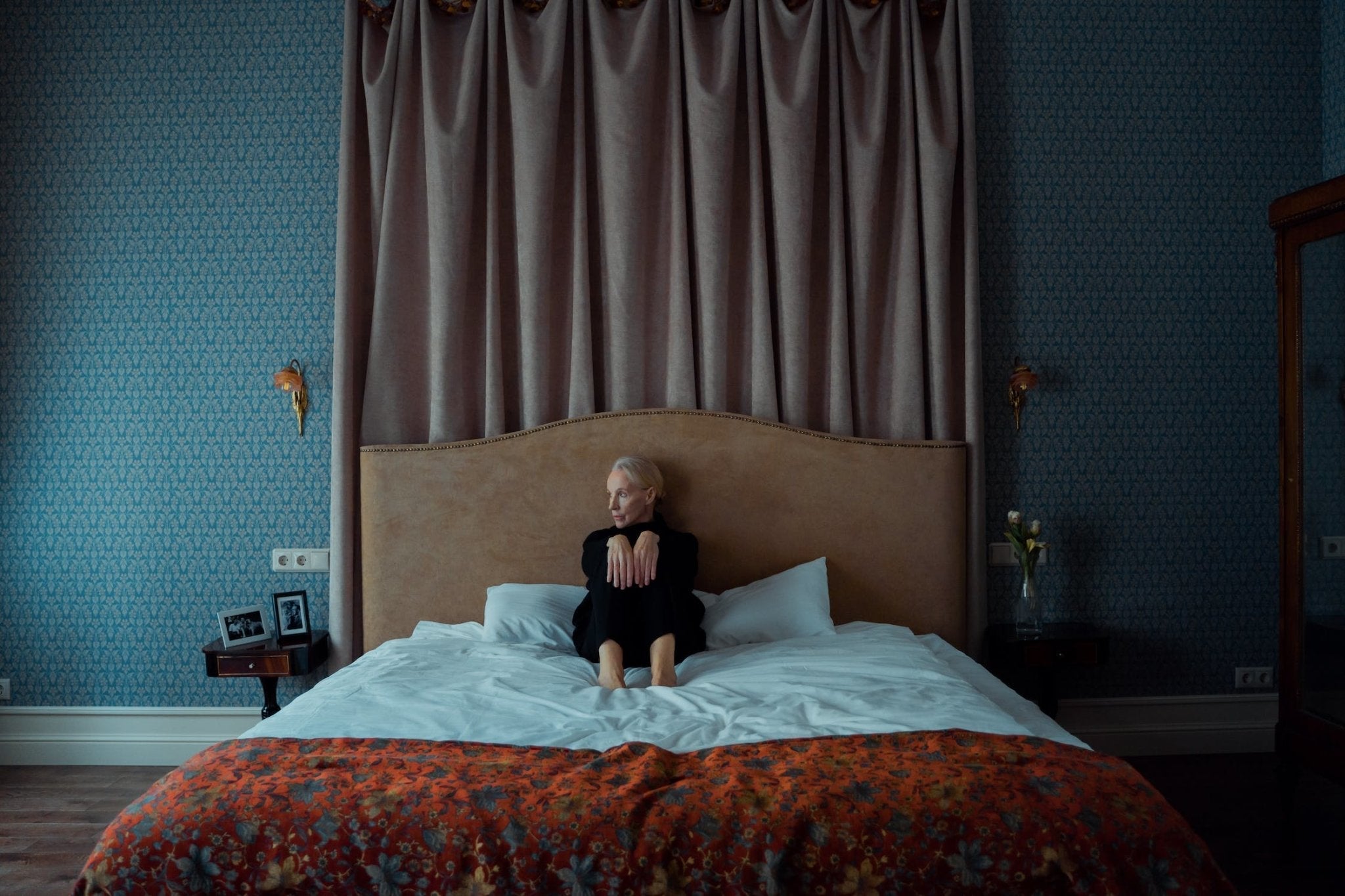Woman sitting on a bed in a cozy room with blue wallpaper and floral decor.