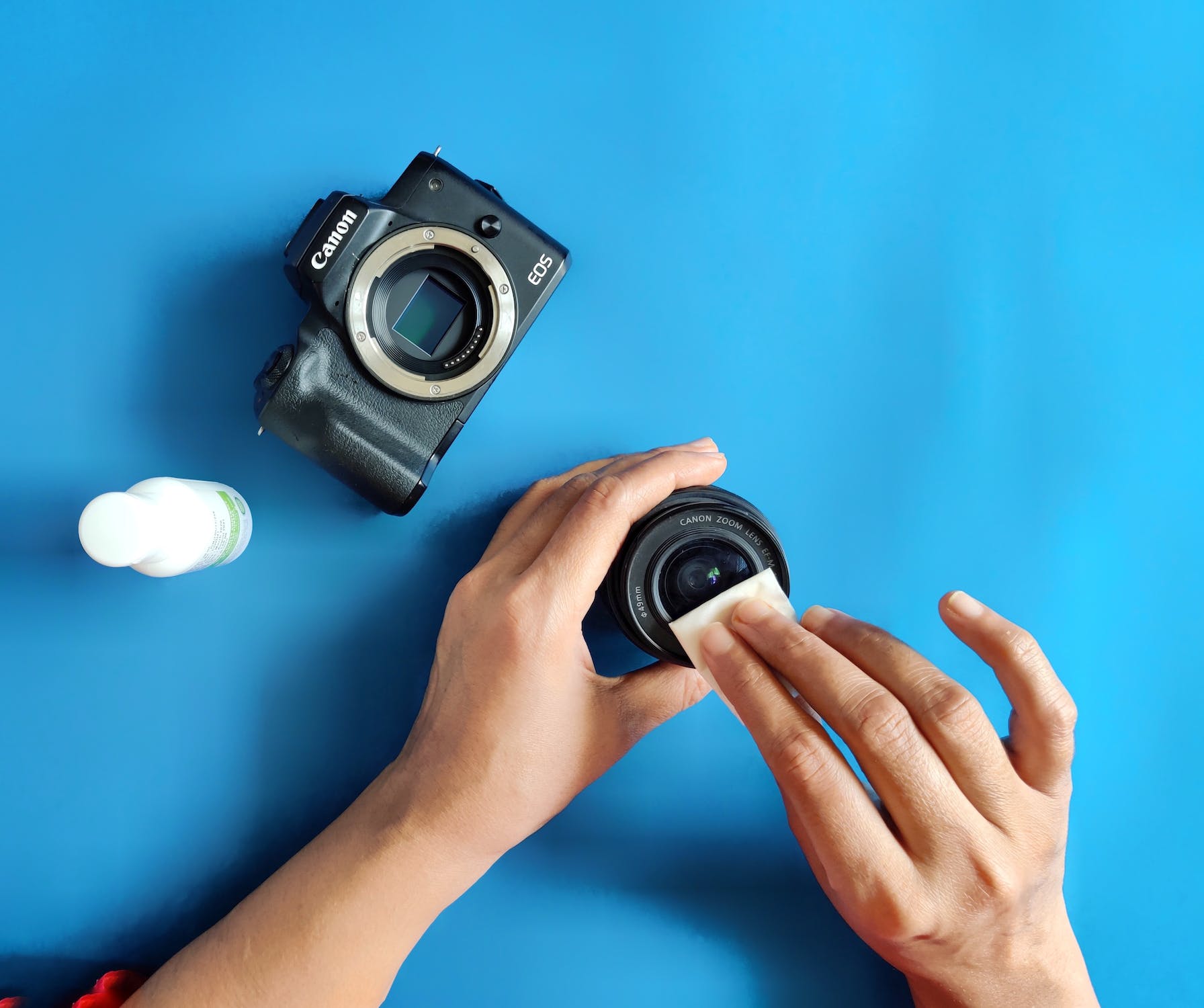 black Canon mirrorless camera with hands cleaning a camera lens and a bottle of cleaning solution on a blue background