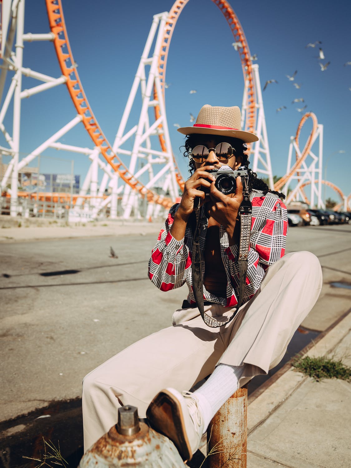 Black man in stylish outfit holding camera near amusement park roller coaster.