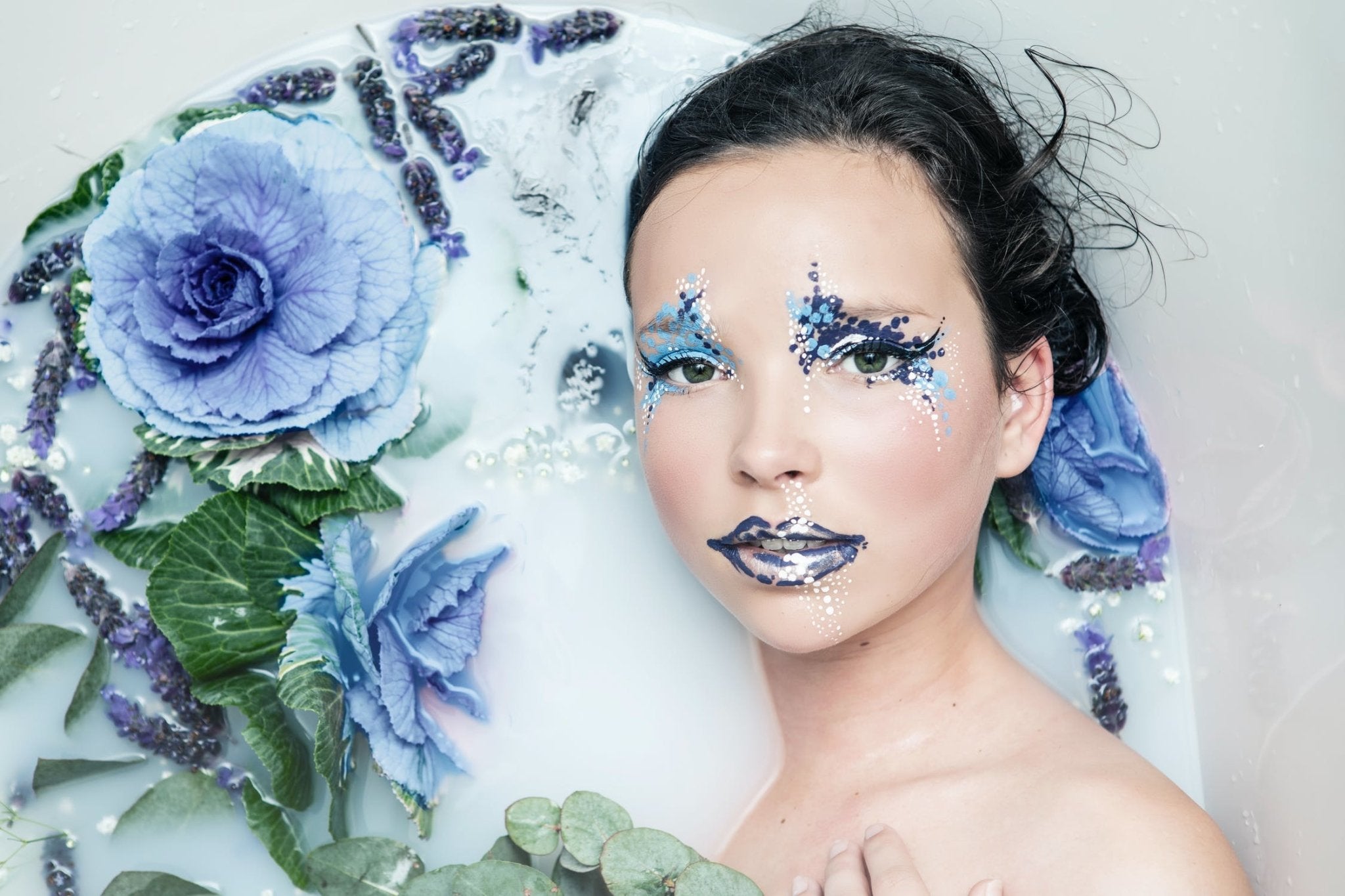 Model with blue floral makeup submerged in milk bath with blue and green flowers around her.