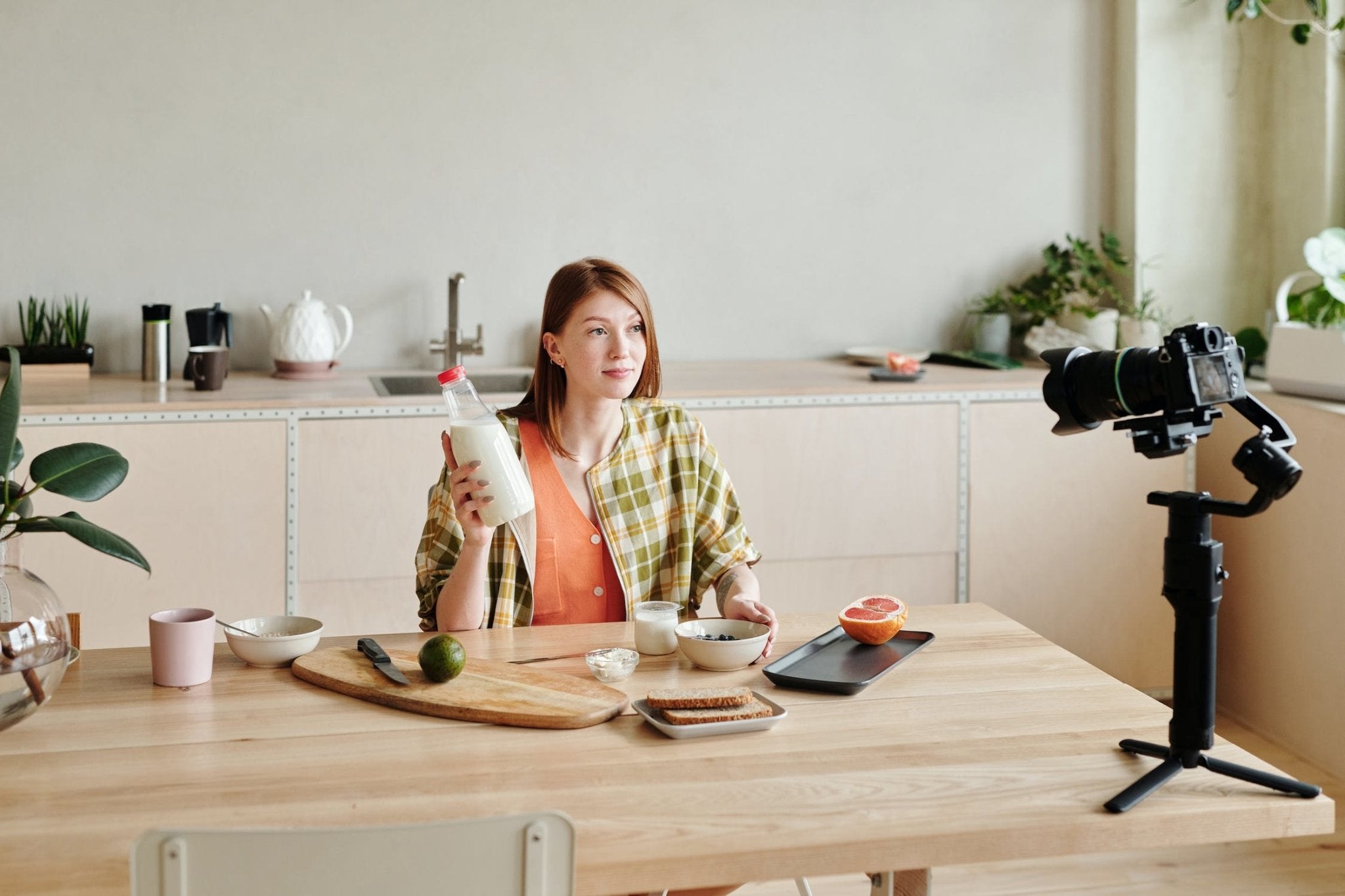 Woman sitting at a kitchen table holding a milk bottle, with a camera setup and breakfast items.