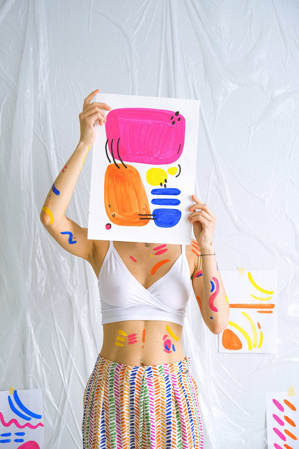 Woman holding a colorful artwork with painted arms, surrounded by abstract art on a white backdrop.