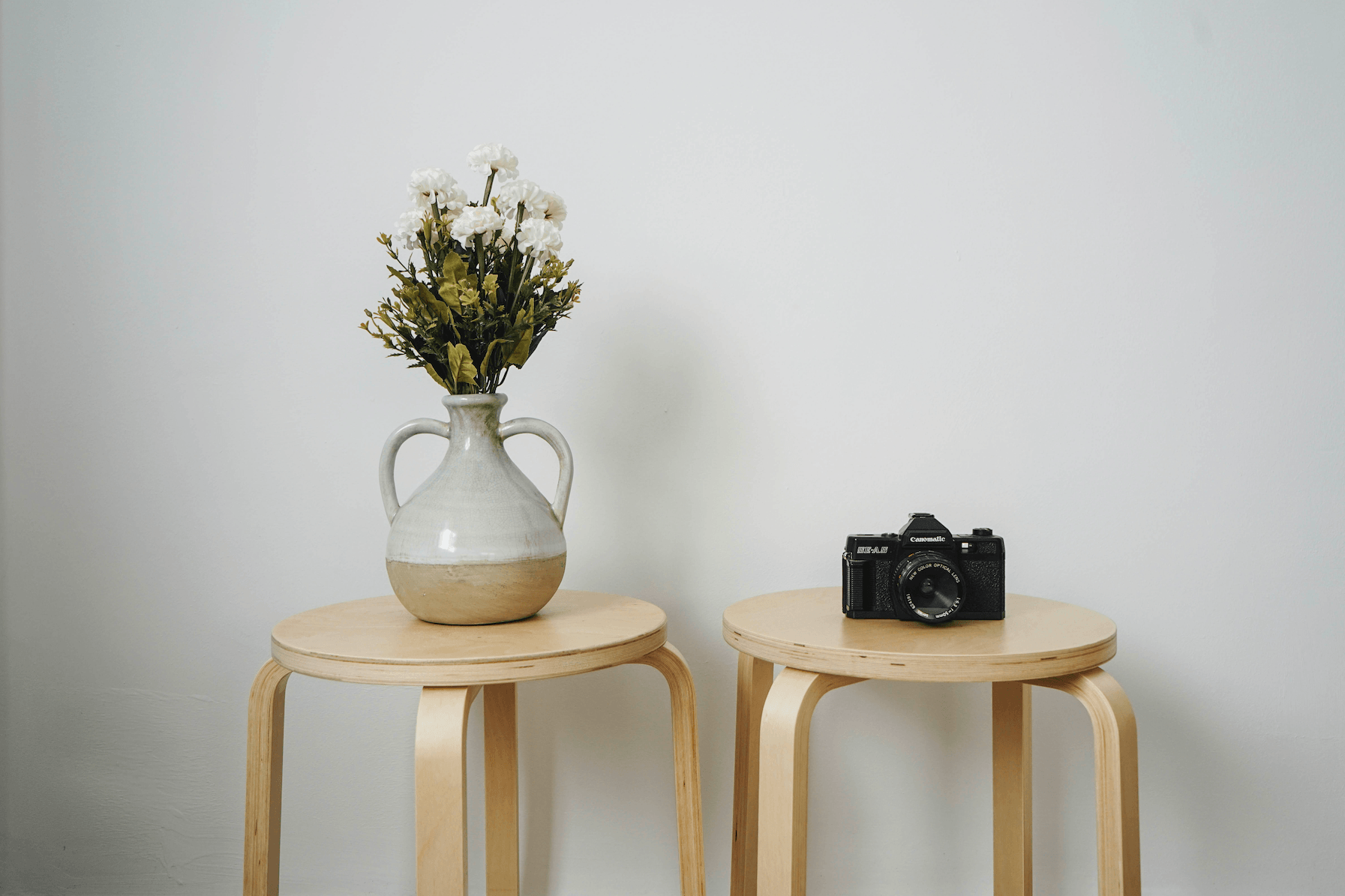 White vase with flowers beside a camera on wooden stools against a white wall
