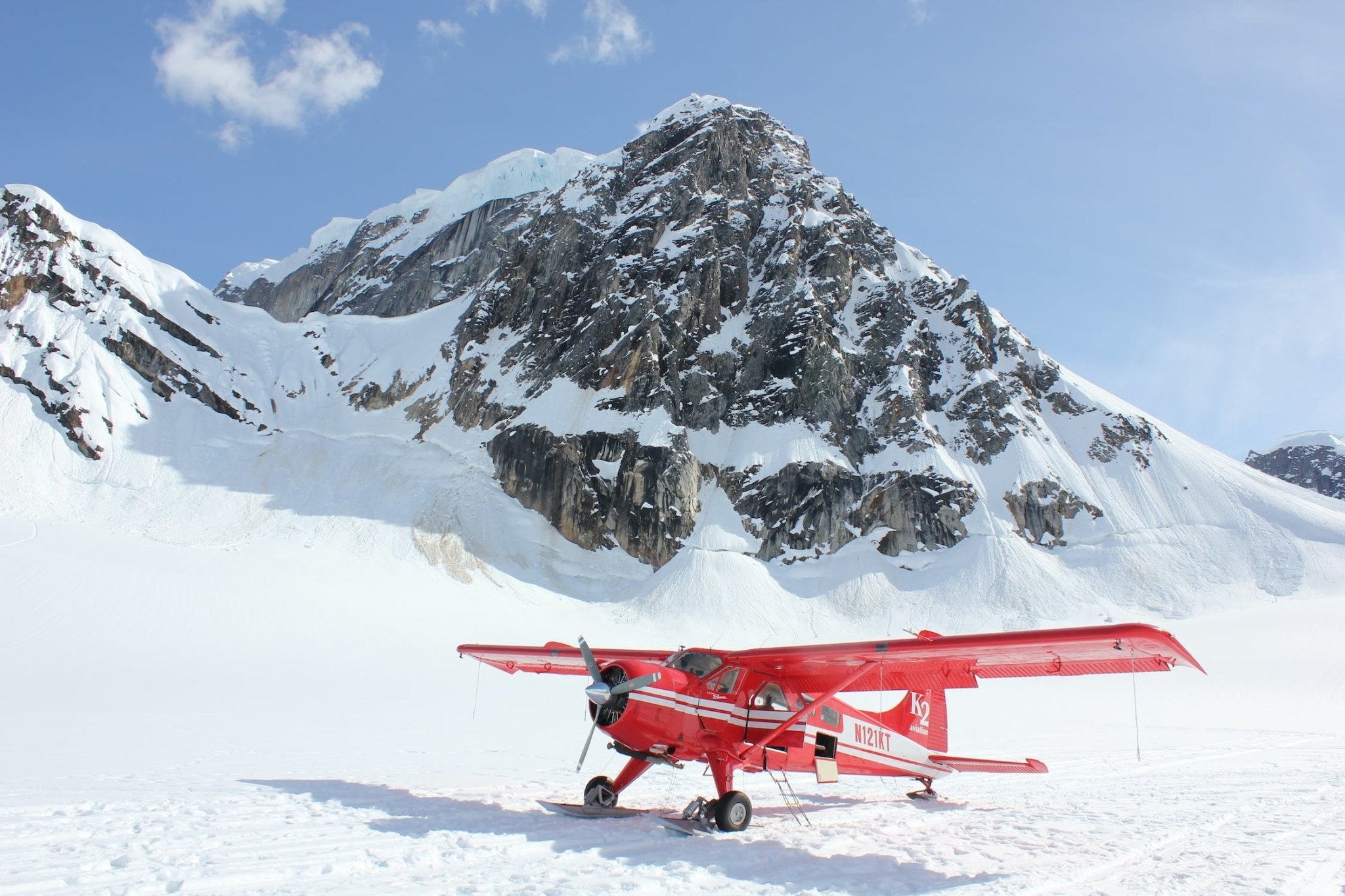 Red plane parked on snow with a mountain backdrop and clear blue sky.