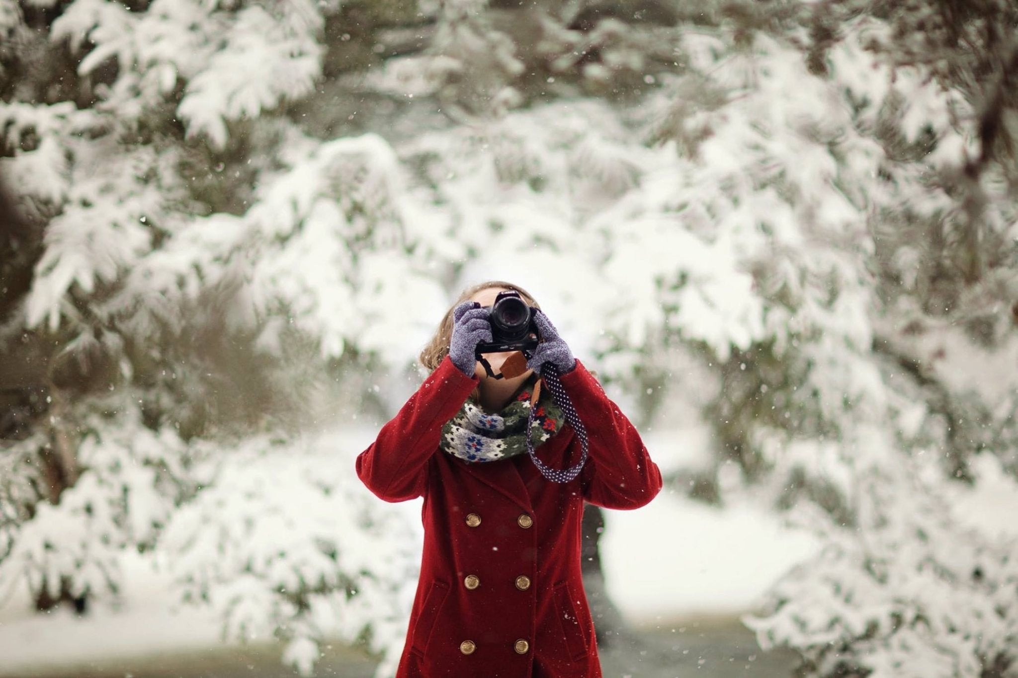 Woman in a red coat photographing snowflakes with a camera amidst falling snow.