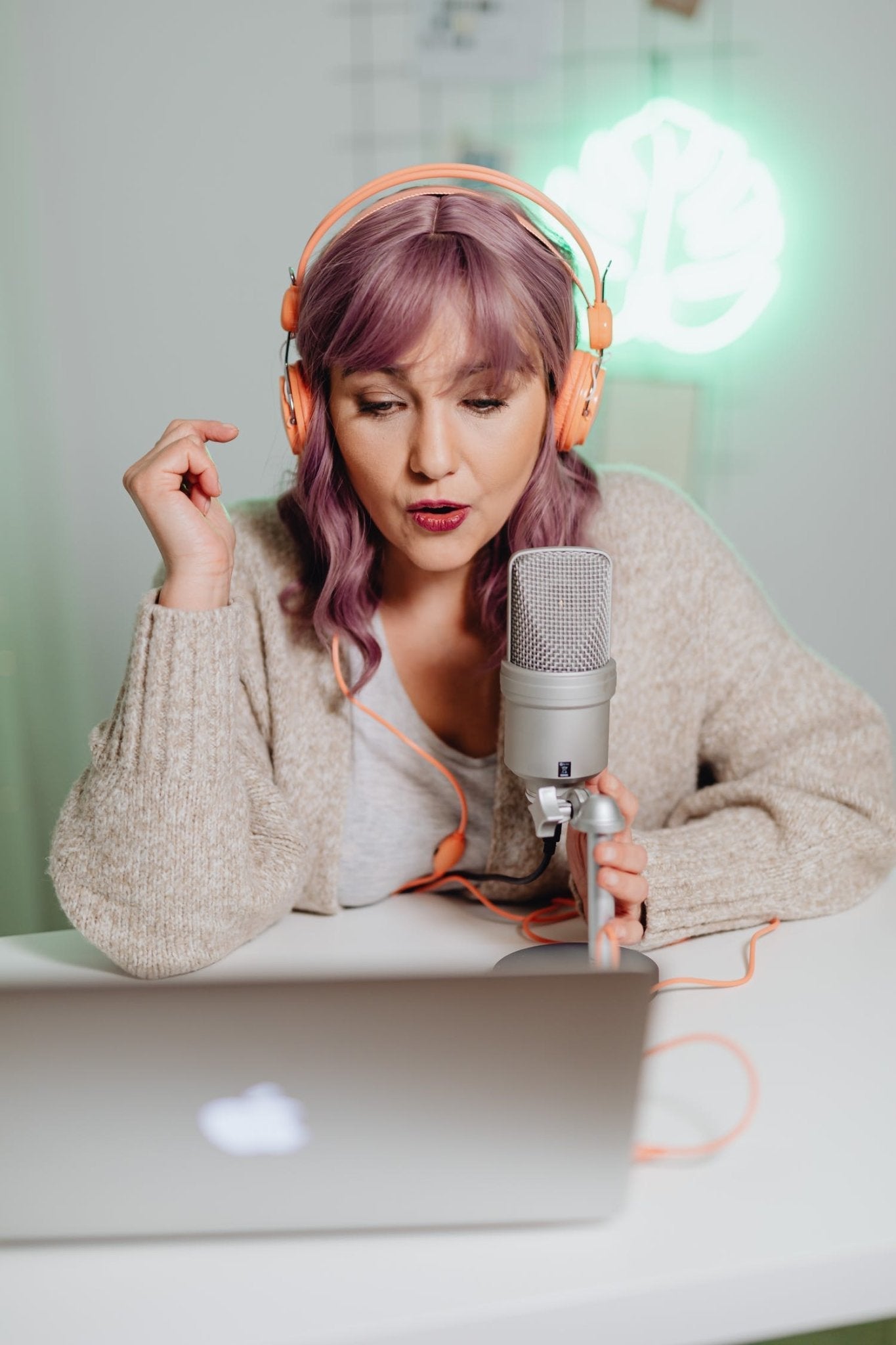 Woman with pink hair wearing headphones, speaking into a microphone, sitting at a laptop on a desk.