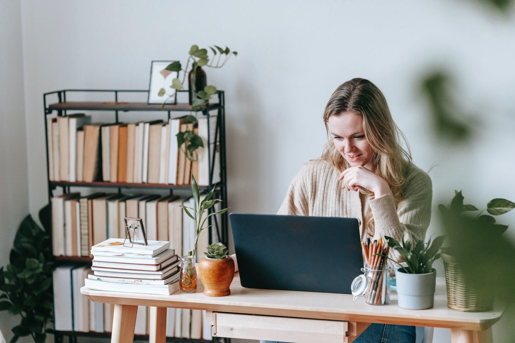 Woman working on a laptop at a home office desk surrounded by plants and books.