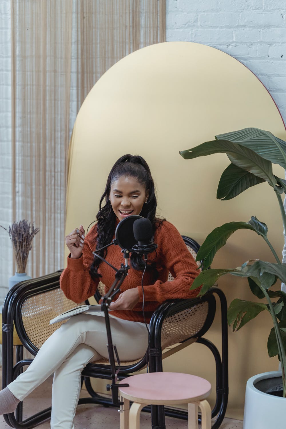 Woman with long hair sitting in front of a microphone, smiling and holding a pen, surrounded by plants.