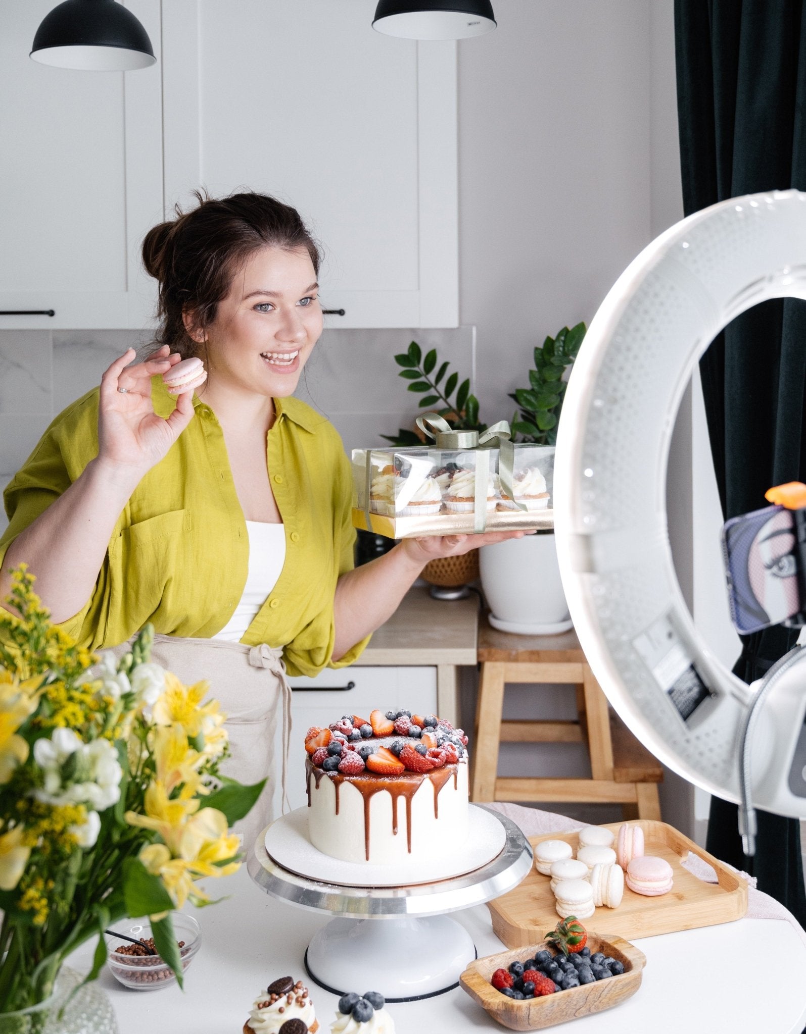 Woman in green shirt holding dessert tray with cake and macarons, surrounded by flowers and a ring light.