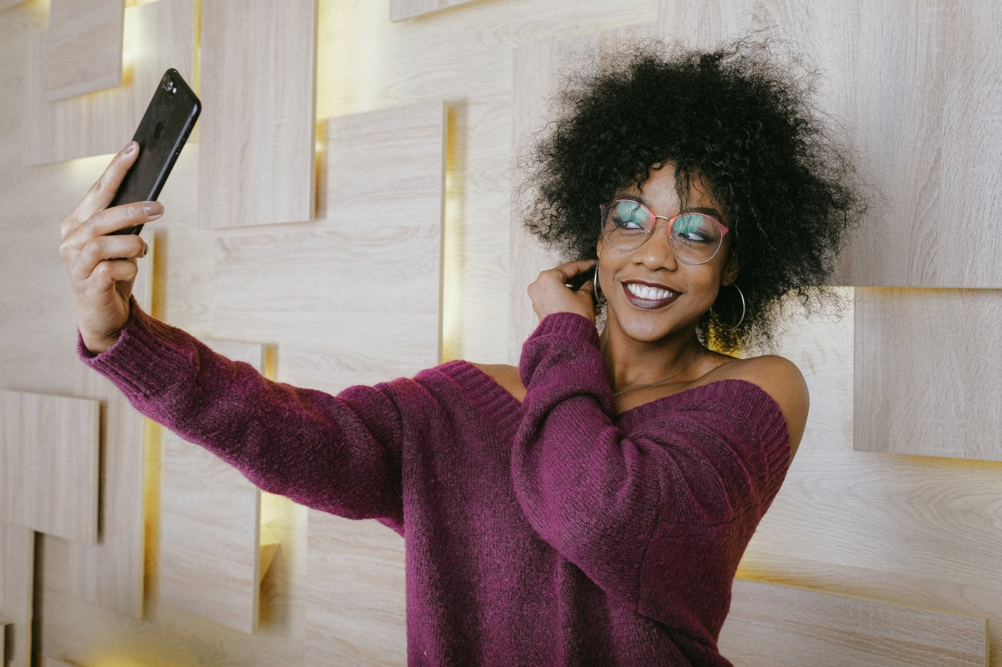 Brown with purple sweater woman taking a selfie with glasses and curly hair, smiling against a wooden background.