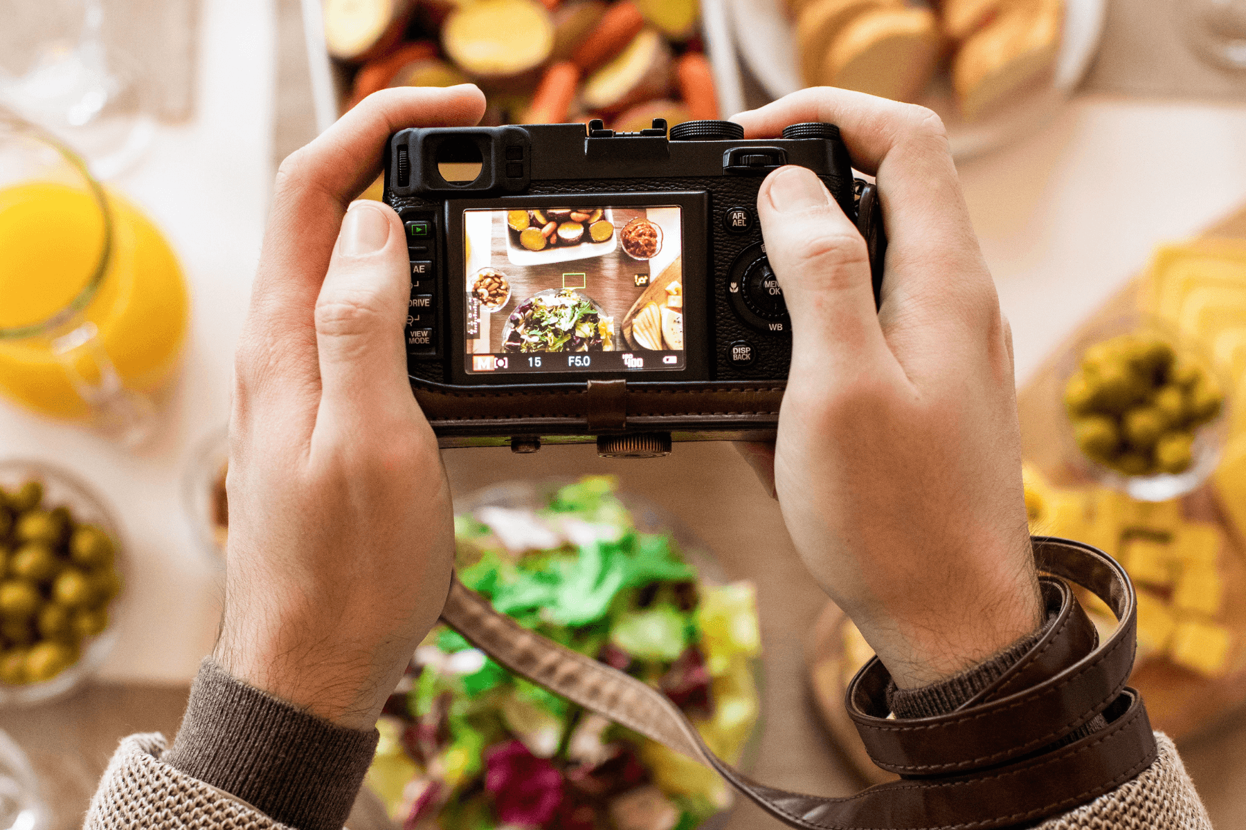 hands holding a camera capturing a beautifully arranged table of food with salad, fruits, and drinks
