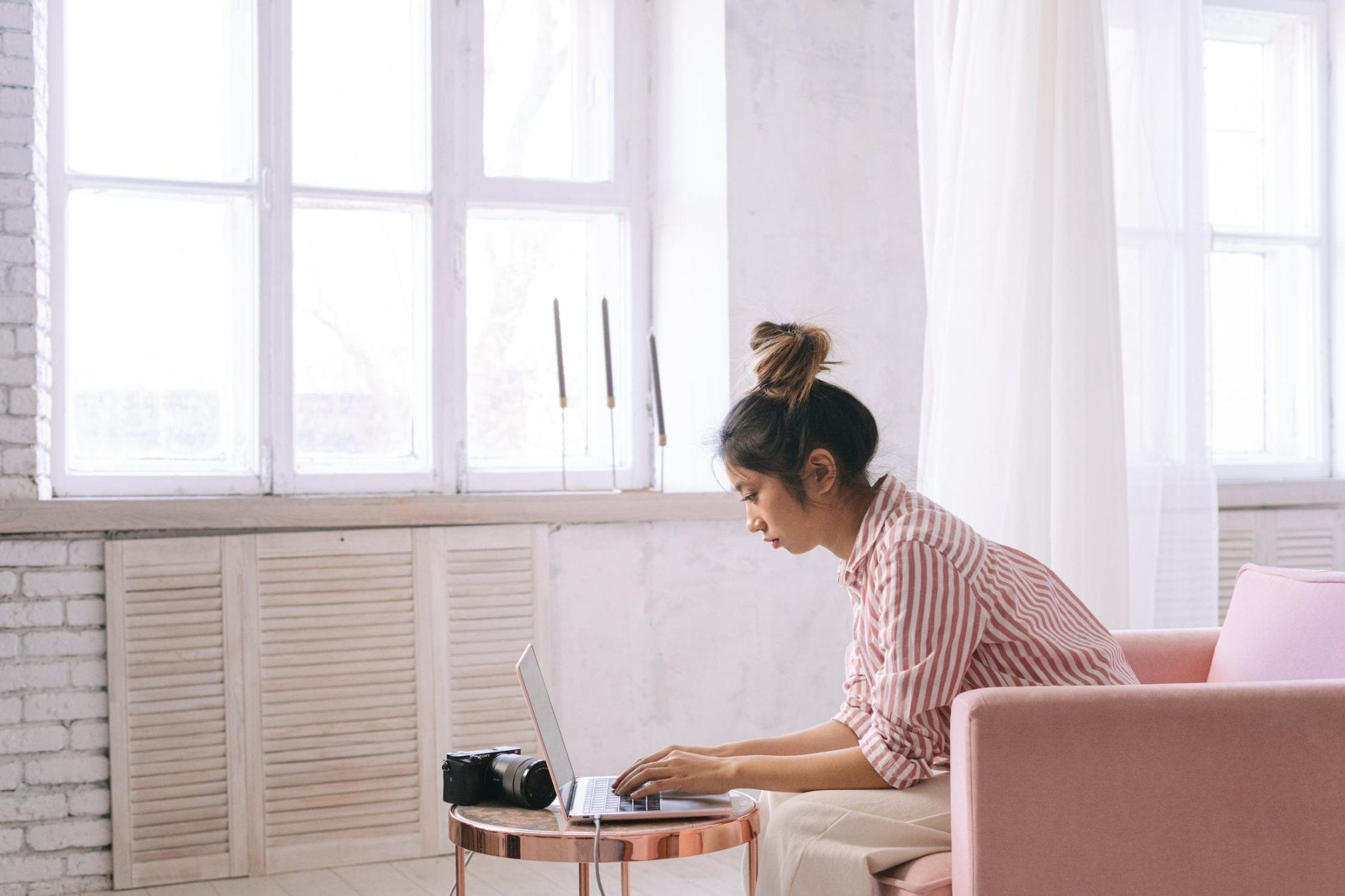 A woman sitting on a pink couch, using a laptop in a bright, airy indoor space.
