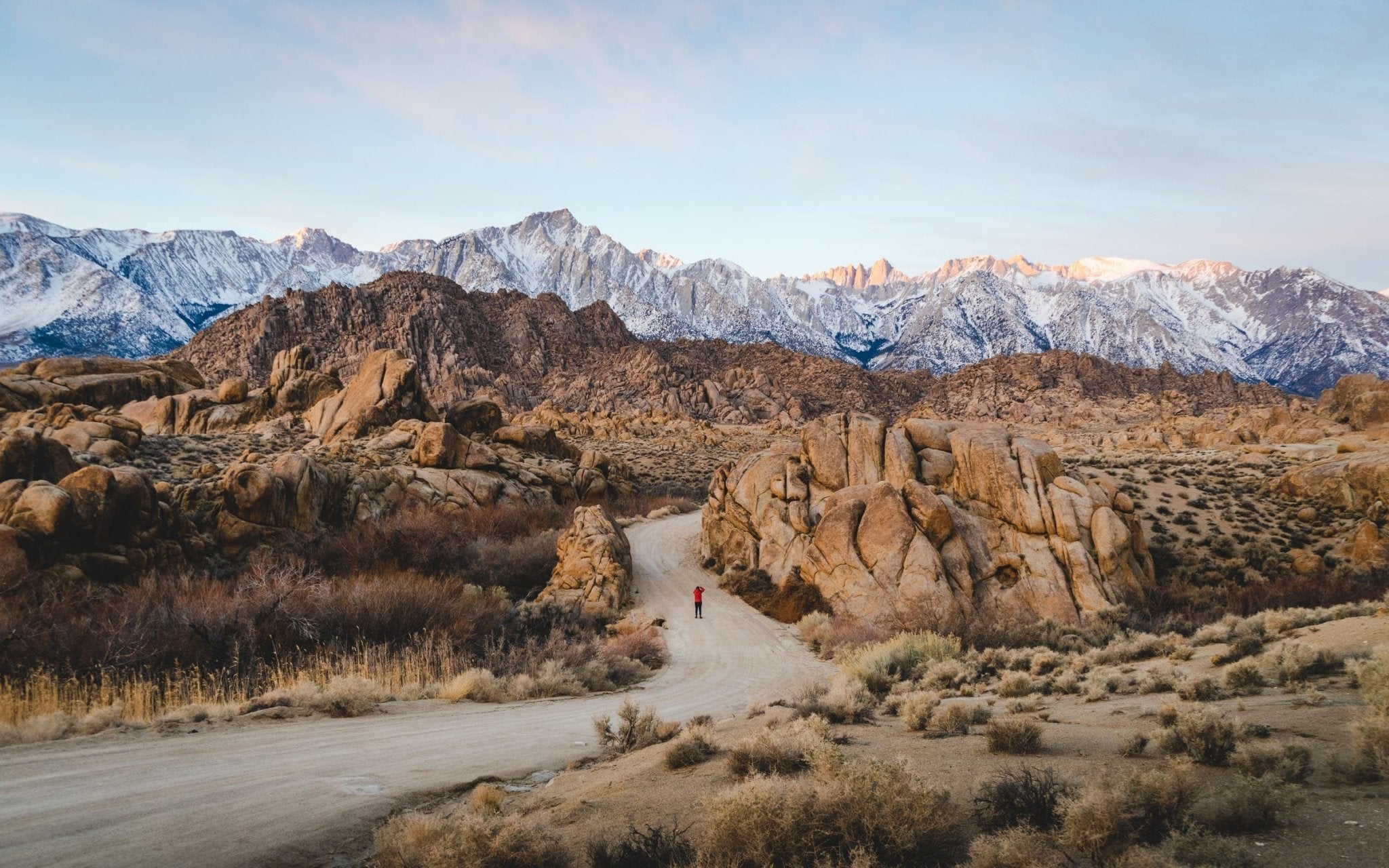 Red hiking figure on a winding dirt path surrounded by rocky mountains and sparse vegetation.