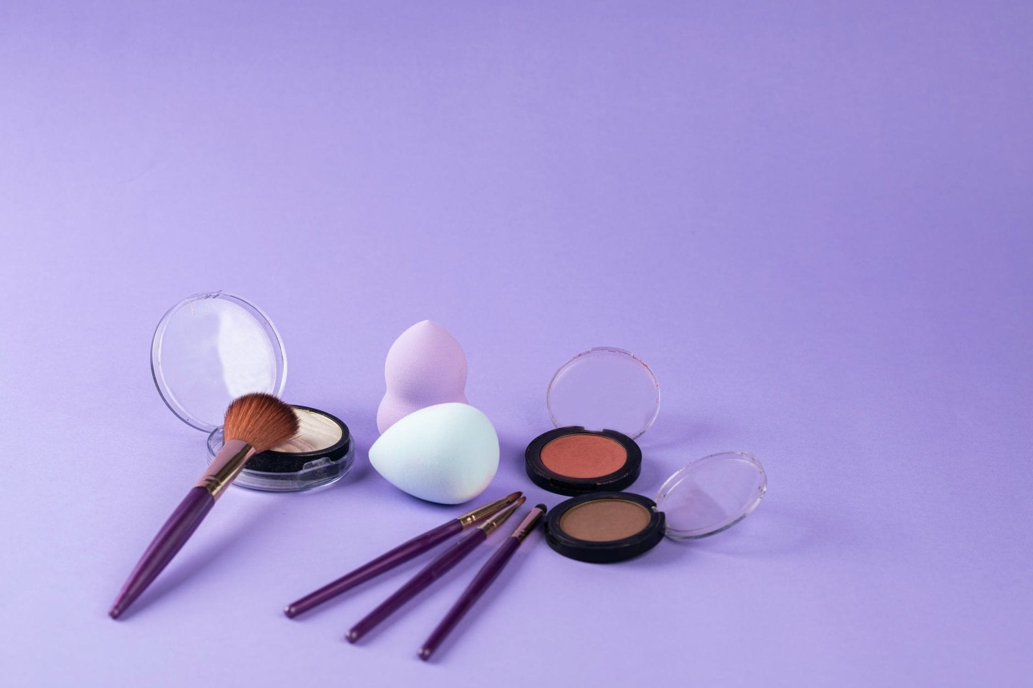 Purple makeup brushes, sponges, and powder compacts on a lilac background.