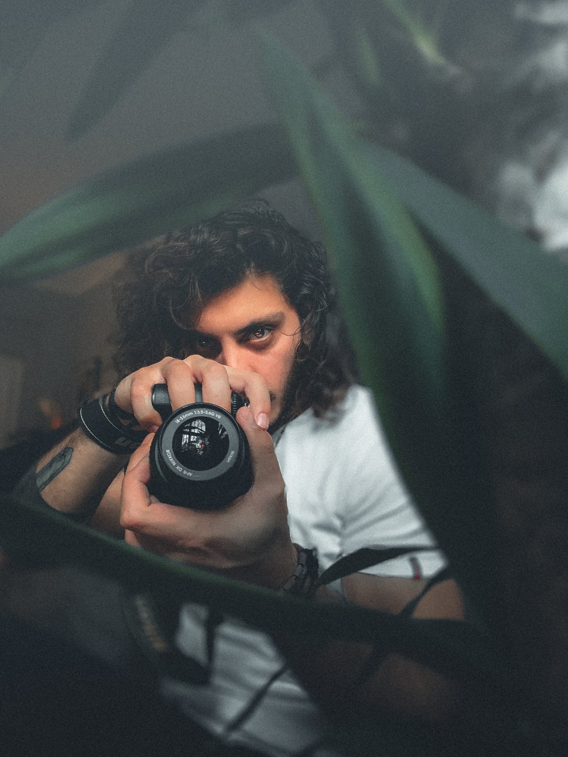 Person holding a camera, partially obscured by plant leaves, with a focused expression.