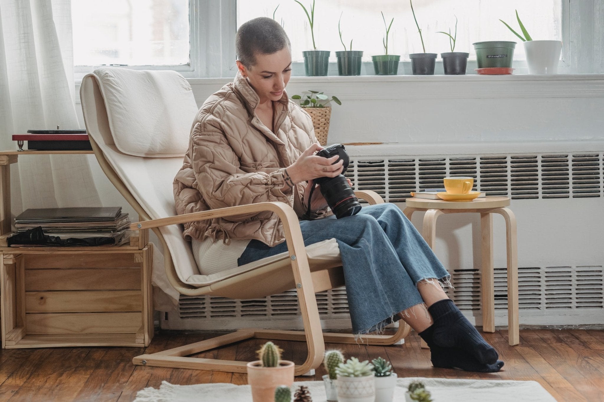 Woman in a quilted jacket sitting in a chair, examining a camera with plants and a cup nearby.