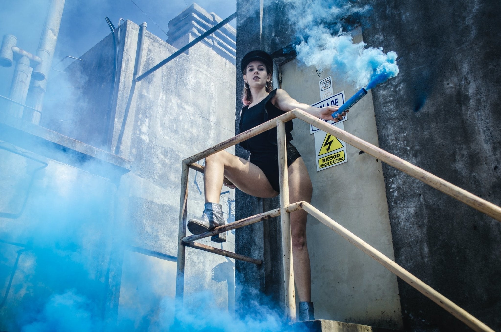 A woman in a black outfit holds a blue smoke bomb while posing on a staircase.