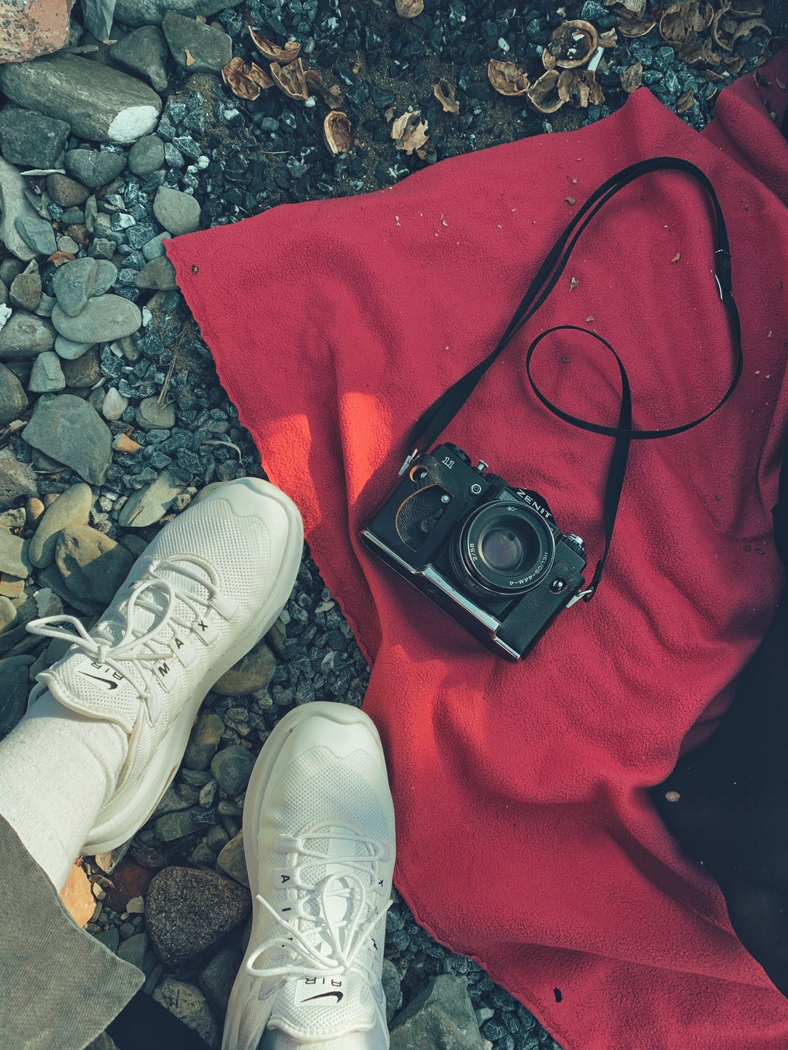 White sneakers and a vintage camera on a red blanket among stones and dried leaves.