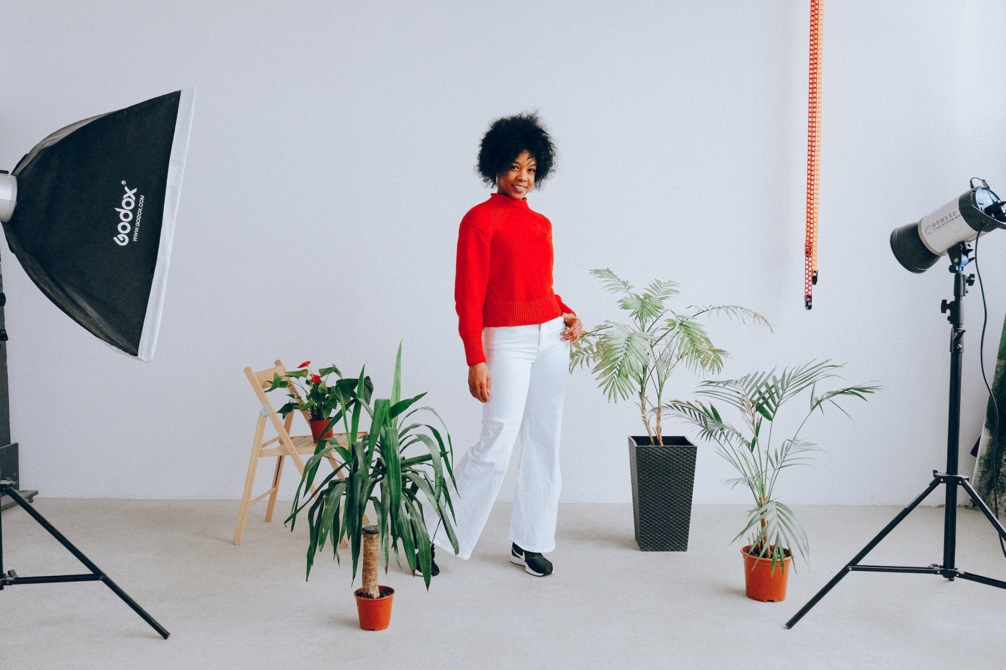 Woman in a red sweater and white pants stands among potted plants in a photography studio.