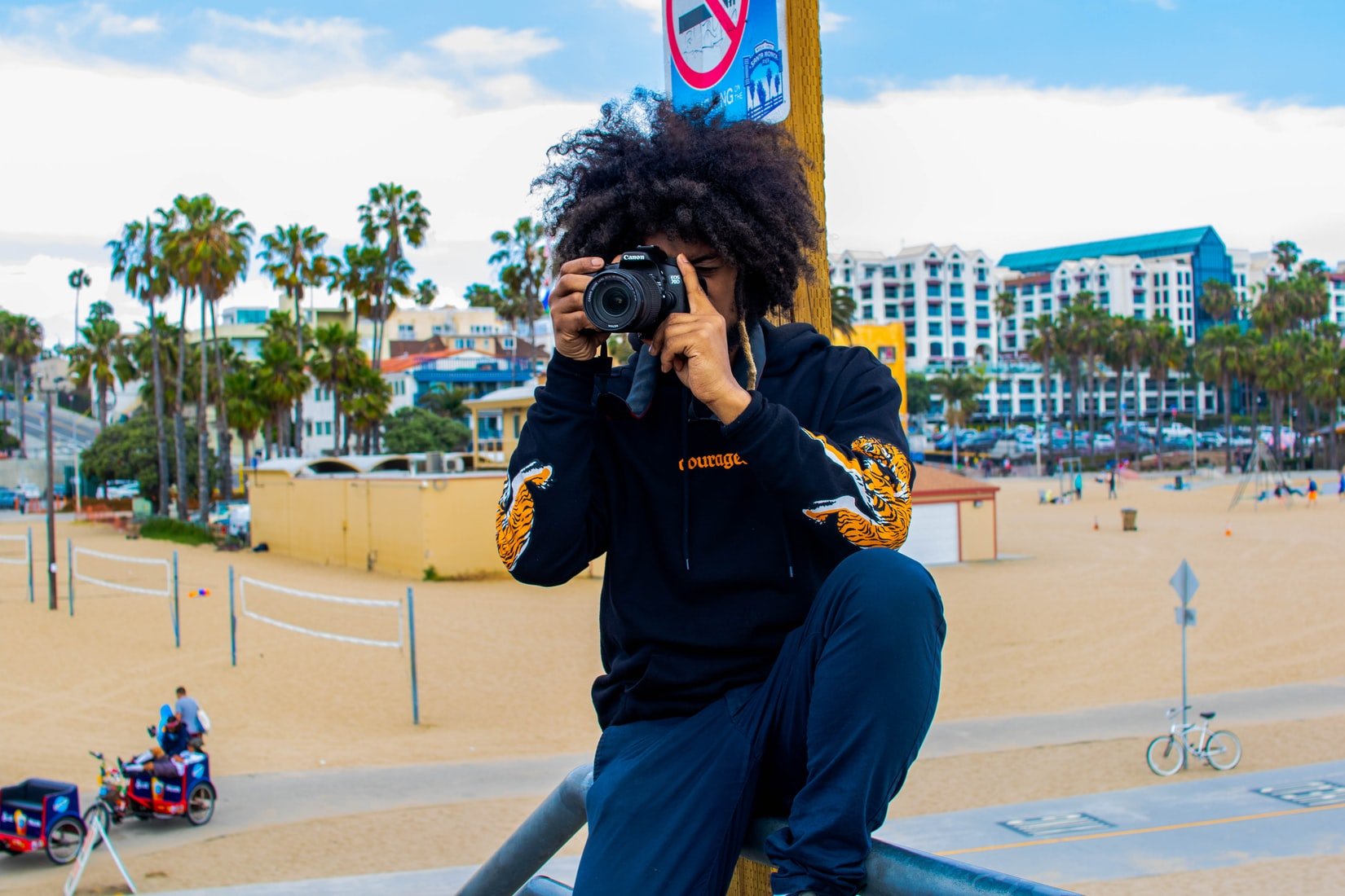 Person with curly hair holding a camera, wearing a black hoodie, near a beach with palm trees.