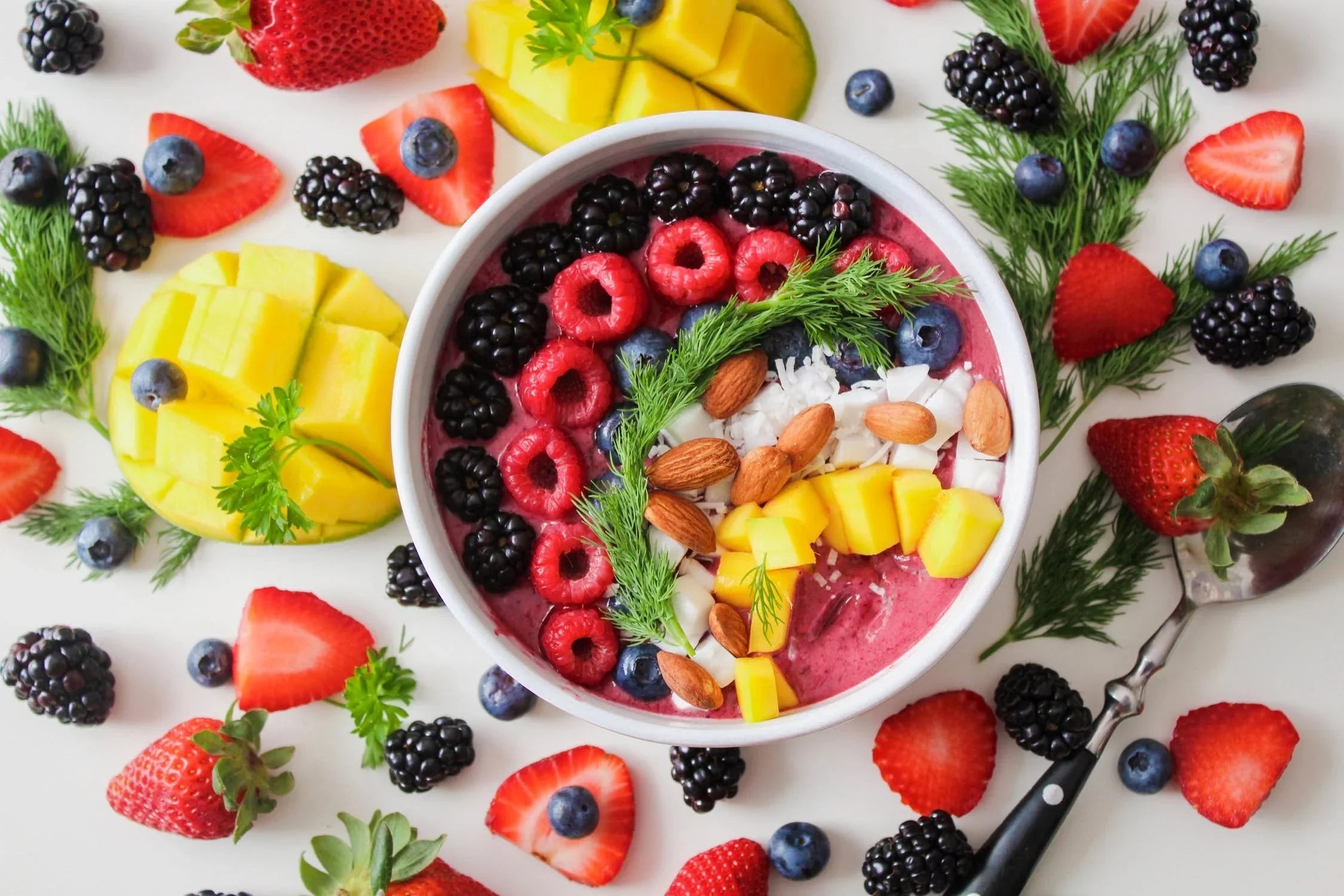 Colorful fruit smoothie bowl topped with berries, mango, almonds, and herbs, surrounded by fresh fruit.