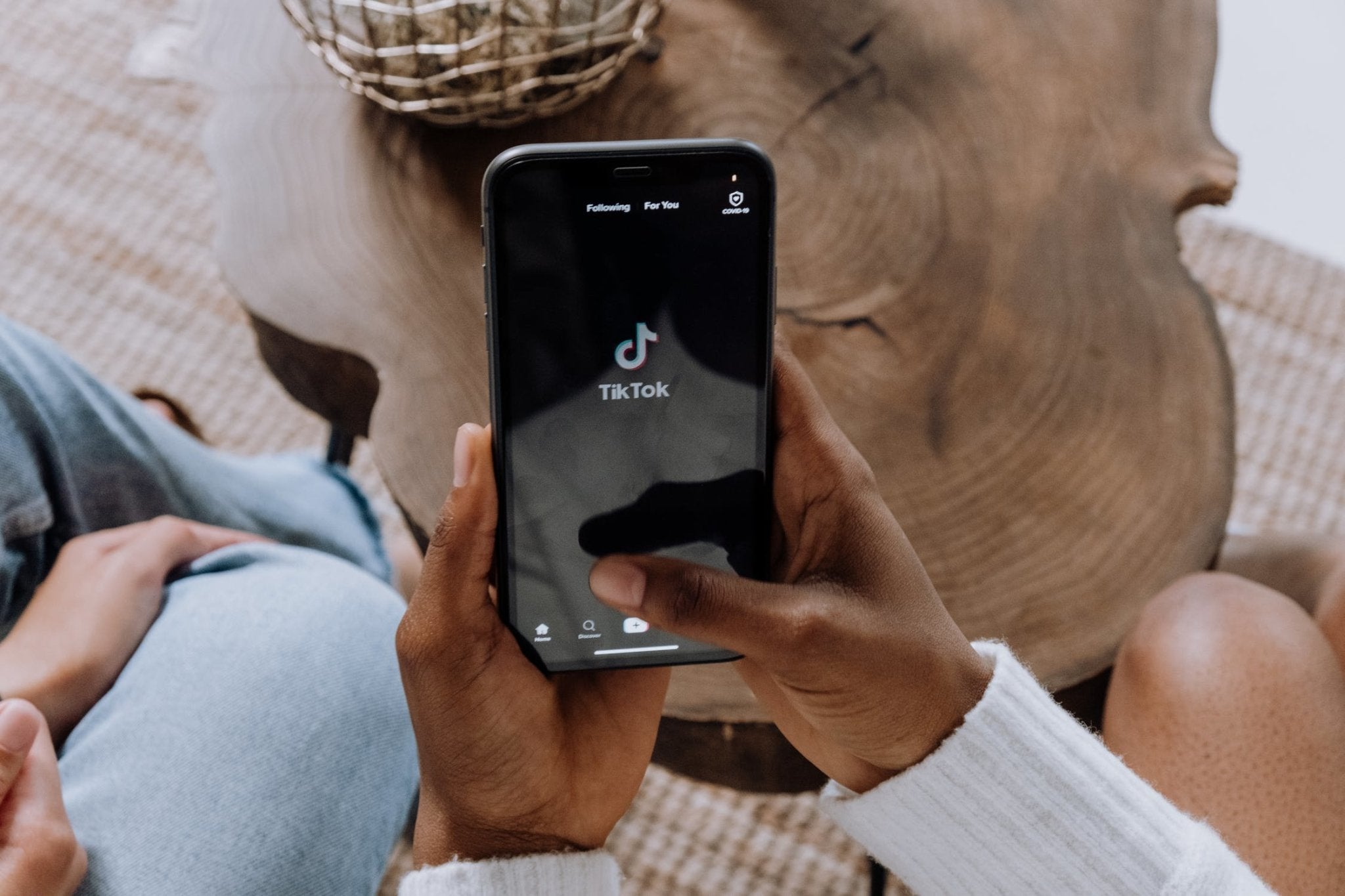 hands holding a smartphone showing the TikTok logo on the screen, placed on a wooden table