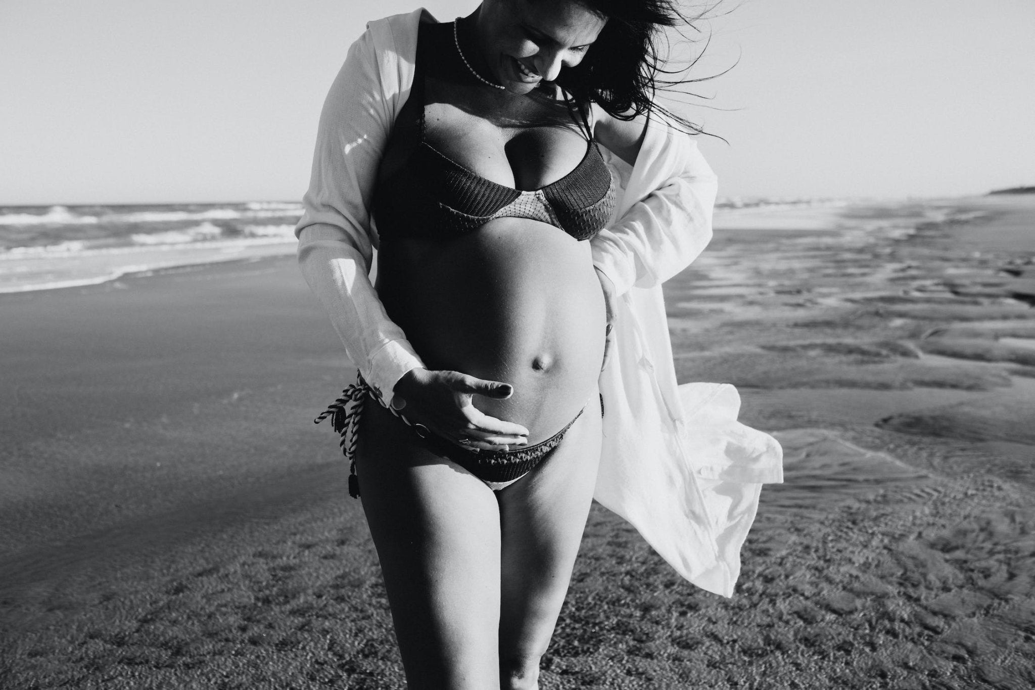 Pregnant woman in a bikini holding her belly while walking on the beach at sunset.