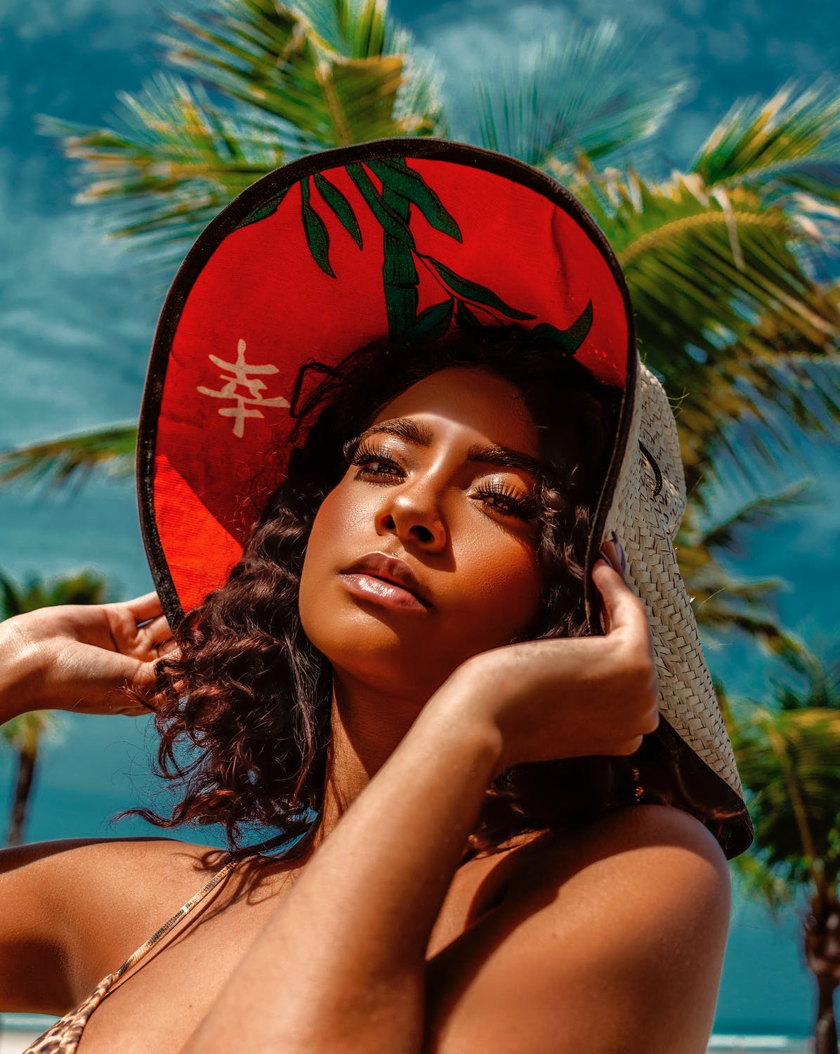 Young woman with curly hair wearing a wide-brimmed hat surrounded by tropical palm trees.