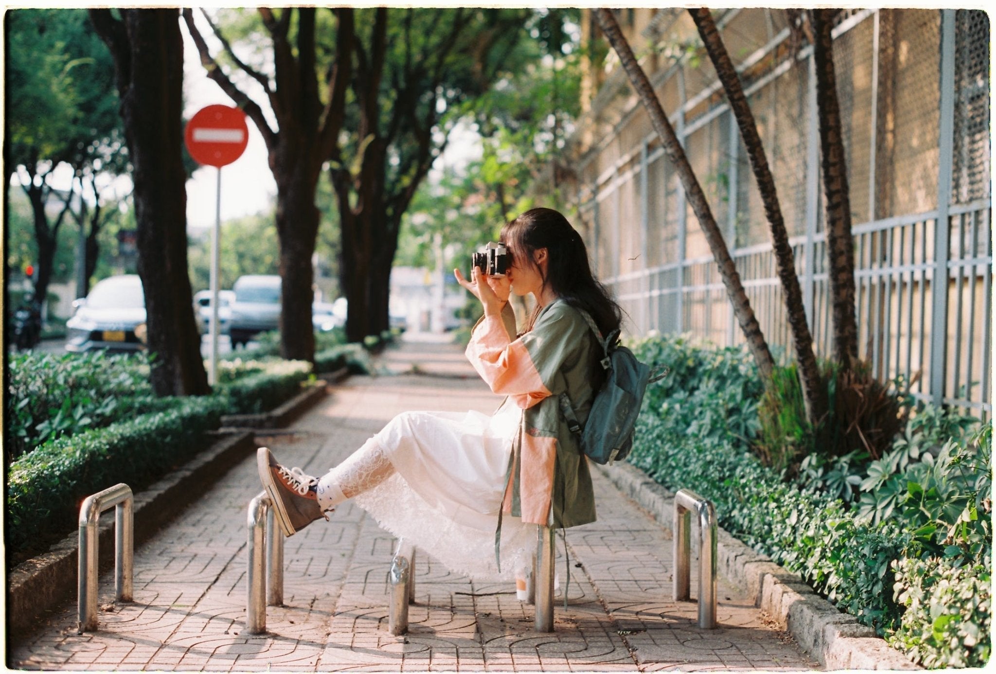 Woman with camera sitting on a sidewalk surrounded by trees and greenery.