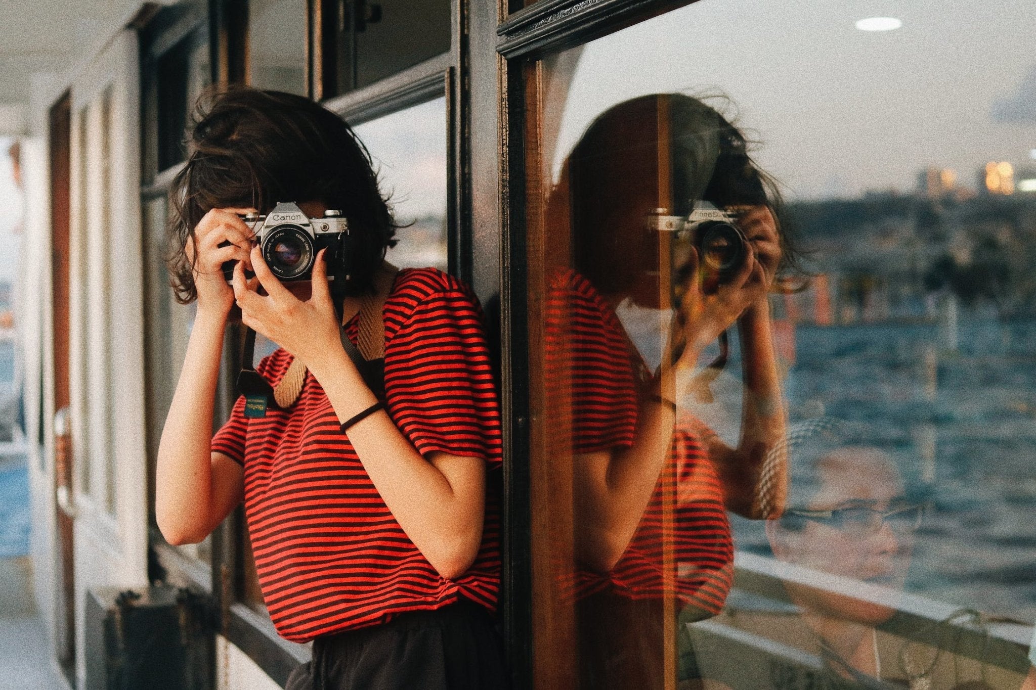 Person in a striped shirt taking a photograph, reflection visible in a window.