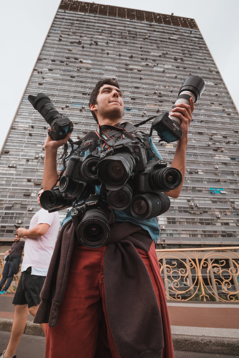 A person holding multiple cameras and lenses against a city building backdrop.